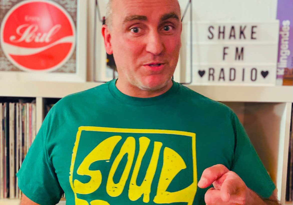 Soulbrother Ali schaut in die Kamera und zeigt mit einem Finger auf die Kamera. Im Hintergrund erkennt man ein Plattenregal und einen Schriftzug Shake!FM Radio