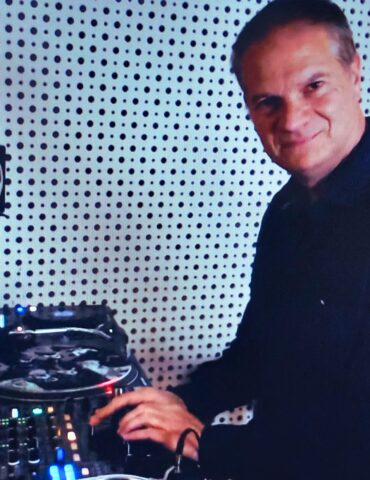 Funky Stevie schaut in die Kamera während er vor einem DJ Mixing Deck steht in einem Radiostudio steht.