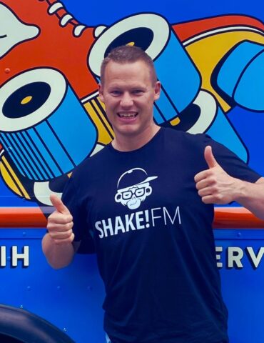 Hajo steht vor einem ANhönger mit Shake!FM T-Shirt, lächelt und streckt beide Daumen nach oben