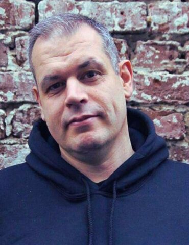 Udo Sievering im Porträt lächelnd steht er vor einer Mauer